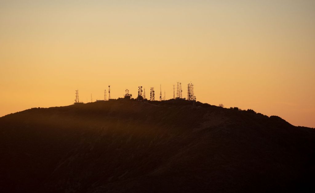 Silhouetted broadcast transmitter antennas on a hilltop against an orange sunset sky