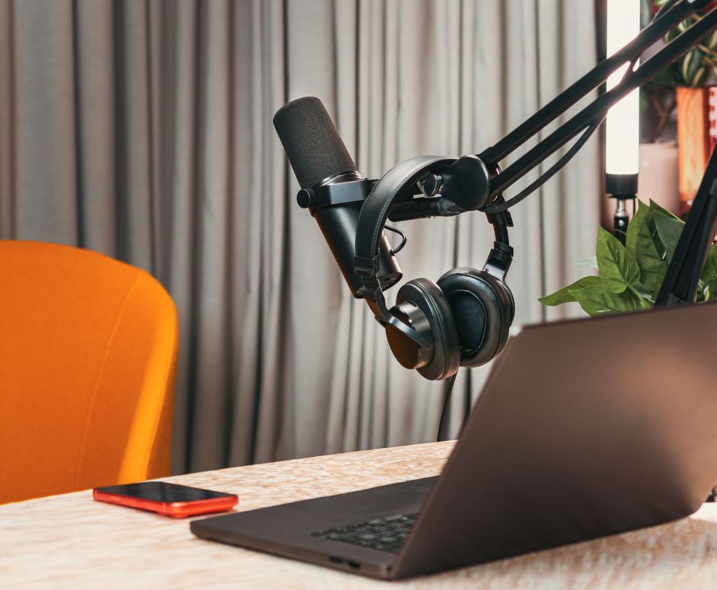 Headphones hanging on a podcast microphone above a laptop and smartphone