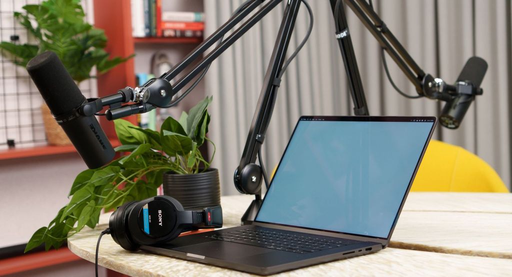 Two podcast microphones on boom arms beside an open laptop on a desk