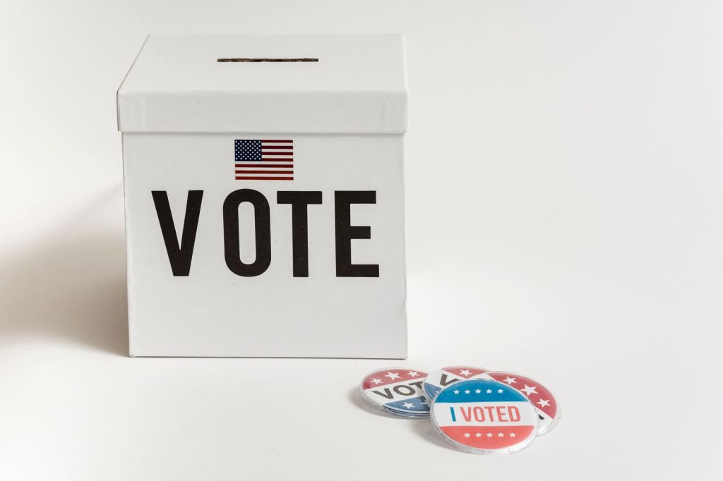 White ballot box marked VOTE with a small American flag, next to I Voted buttons